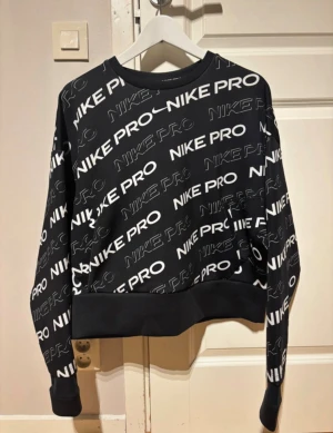 Nike Pro svart sweatshirt med logga - Svart Nike Pro sweatshirt med vitt all-over logomönster. Tröjan har rund hals, långa ärmar och breda muddar vid ärmslut och midja. Tillverkad i Dri-FIT polyester som andas, perfekt för träning eller chill. Snygg och sportig vibe.