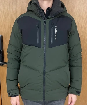 Grön dunjacka från Sail Racing - Snygg grön och svart dunjacka från Sail Racing med huva och flera dragkedjeförsedda fickor. Jackan har en sportig look med logga på bröstet och patch på ärmen. Perfekt för kalla dagar med sin vadderade design och höga krage.