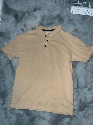 Beige pikétröja från Sams - Snygg beige pikétröja från Samsoe Samsoe med klassisk krage och tre svarta knappar framtill. Tröjan är kortärmad och gjord i mjuk bomull, perfekt för en chill och stilren look. Passar dig som gillar enkel och clean stil.