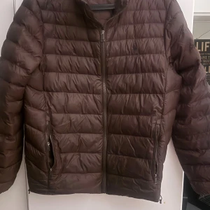 Brun pufferjacka från Polo Ralph Lauren - Säljer en brun pufferjacka från Polo Ralph Lauren med klassisk quiltad design. Jackan har dragkedja framtill, två sidofickor med dragkedja och hög krage. Materialet är glansigt och lätt, perfekt för kyliga dagar. Diskret logga på bröstet. Nypris 4295kr
