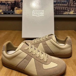 Maison Margiela beige sneakers - Maison Margiela sneakers i en clean beige och krämvit färg. Skorna har en klassisk låg siluett med paneler i mocka och skinn, samt matchande snörning. Perfekta för dig som gillar minimalistisk och stilren design med exklusiv känsla.