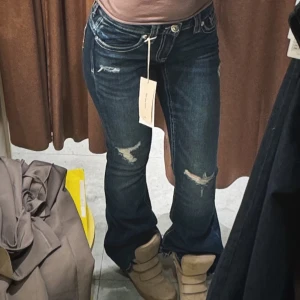 Blå bootcut jeans med slitningar - Snygga mörkblå jeans med bootcut passform och slitna detaljer på låren och knäna. Jeansen har klassisk femficksdesign, låg midja och är tillverkade i jeansmaterial med stretch för extra komfort. Perfekta för dig som gillar en avslappnad och trendig look.