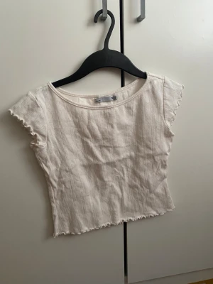 Vit ribbad topp från 157 XS - Säljer en söt vit topp från 157 i storlek XS. Toppen har ribbad struktur med diskreta vertikala mönster, korta ärmar med vågig kant och rund halsringning. Materialet är mjukt och stretchigt, perfekt för en chill look. Passformen är normal och toppen är croppad.