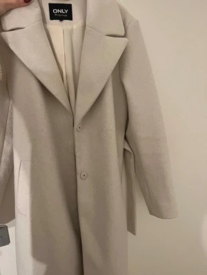 Beige kappa från ONLY med bälte - Stilren beige kappa från ONLY med klassisk krage, knäppning framtill och bälte i midjan. Lång modell med raka ärmar och diskreta fickor. Perfekt för dig som vill ha en clean och tidlös look.