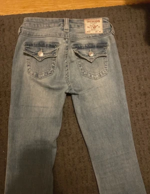 True Religion ljusblå jeans med fickdetaljer - Säljer ett par ljusblå jeans från True Religion med snygga ficklock och silverknappar bak. Jeansen har klassisk femficksdesign och utsvängda ben.