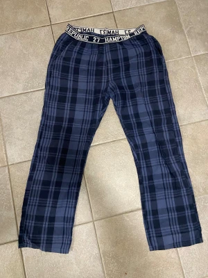 Blårutiga byxor från Hampton Republic - Säljer ett par blårutiga byxor från Hampton Republic med elastisk midja och logotypdetaljer. Byxorna har en avslappnad passform och är gjorda i mjukt bomullsmaterial, perfekta för chill dagar hemma. Snyggt rutmönster i mörkblå toner. Sparsamt använda, så fint skick.