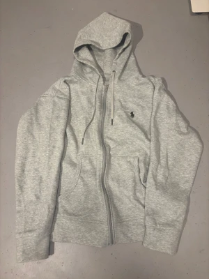 Grå hoodie från Polo Ralph Lauren - Snygg ljusgrå hoodie från Polo Ralph Lauren med dragkedja, huva med snörning och klassisk logga broderad på bröstet. Tröjan har långa ärmar och två fickor framtill. Perfekt för chill dagar och enkel att matcha med jeans eller joggers.