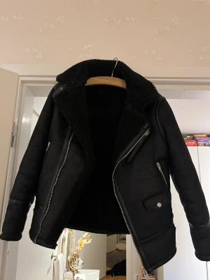 Svart teddyjacka från Zara - Säljer en svart teddyjacka från Zara med mjukt foder, bred krage och detaljer i skinnimitation. Jackan har dragkedjor, fickor och bältesdetaljer nertill. Perfekt för dig som vill ha en snygg och varm jacka med cool bikerstil.