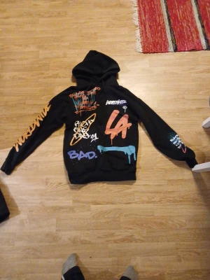 Svart hoodie Black Squad XS graffiti - Svart hoodie från Black Squad i storlek XS med relaxed fit. Tröjan har färgglada graffiti-prints och text över hela, snörning vid huvan och lång ärm. Snygga detaljer i orange, blått och vitt som ger streetkänsla. Perfekt för dig som gillar bold stil.