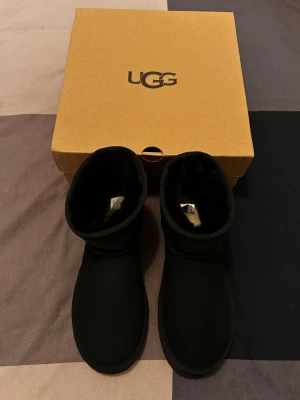 Svarta UGG Classic mini II - Säljer ett par klassiska svarta UGG boots med rund tå och platt sula. Bootsens utsida är i mjuk mocka och insidan är fodrad med fluffigt syntetmaterial för extra värme. Perfekt för kalla dagar och har en enkel, stilren design.