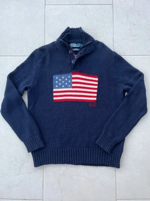 Polo Ralph Lauren USA Iconic sweater - Mörkblå stickad tröja från Polo Ralph Lauren med amerikansk flagga framtill. Tröjan har hög krage med både dragkedja och knappar, samt ribbade muddar vid ärmslut och nederkant. Materialet är 100% bomull och modellen är långärmad.  (Nypris cirka 5000kr).                  Mått📏 Längd: 71cm Bröstbredd:55cm Ärmlängd:68cm 