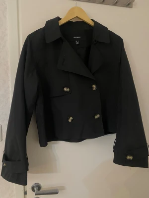 Trenchcoat - Svart trenchcoat från Vero Moda med dubbelknäppning och klassisk krage. Storlek M men passar även S. Jackan är croppad och har snygga detaljer med knappar på ärmsluten. Inga defekter. Endast använt fåtal gånger💞