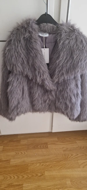 Grå pälsjacka från Design by Amor - Säljer en oanvänd fluffig och trendig grå pälsjacka från Design by Amor. Jackan har lång lugg, bred krage och är helfodrad för extra komfort. Nypris 600 kr