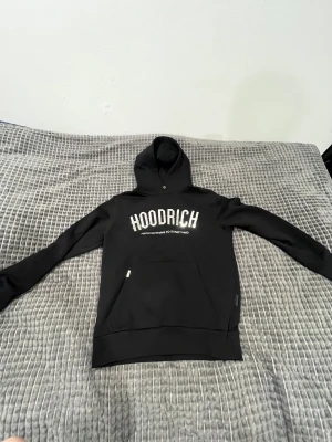 Svart Hoodie från Hoodrich S - Svart hoodie från Hoodrich med stor, glansig silvertext på bröstet och texten 'FROM NOTHING TO SOMETHING' under. Klassisk känguruficka framtill, huva med knappdetalj och långa ärmar. Perfekt för dig som gillar streetwear och vill sticka ut.