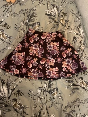 Blommig kortkjol från Soul River XS - Supersöt kortkjol från Soul River i storlek XS. Kjolen har ett mörkt tyg med lila och rosa blommor, och en smidig dragkedja bak. Den har en lätt utsvängd form och är gjord i mjuk polyester som känns skön mot huden. Perfekt för dig som gillar romantiska vibes och blommigt!💞