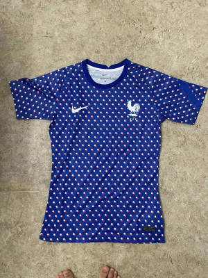 Blå fransk fotbollströja Nike - Säljer en blå fotbollströja från Nike med prickigt mönster i vitt och rött. Tröjan har korta ärmar och rund hals. På bröstet finns Frankrikes landslagsemblem och Nike-logga. Materialet är lätt och andas, perfekt för träning eller match.
