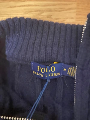 Marinblå stickad tröja Polo Ralph Lauren - Snygg marinblå stickad tröja från Polo Ralph Lauren med halv dragkedja och ribbad krage. Tröjan har ett klassiskt kabelstickat mönster och den ikoniska röda logon broderad på bröstet. Perfekt för dig som gillar stilrena och tidlösa plagg.