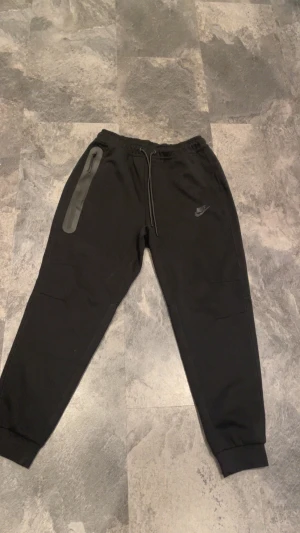 Svarta mjukisbyxor från Nike, storlek L - Svarta mjukisbyxor från Nike i storlek L med elastisk midja och snörning. Byxorna har en modern tapered passform, ribbade muddar vid bensluten och en snygg dragkedja på ena fickan. Perfekta för chill eller träning.