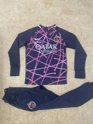PSG långärmad träningströja Nike - Säljer en mörkblå långärmad träningströja från Nike x PSG med coolt rosa och lila grafiskt mönster framtill. Tröjan har PSG-logga och Qatar Airways-tryck på bröstet, samt vita detaljer vid halsen. Perfekt för fotbollsträning eller chill.