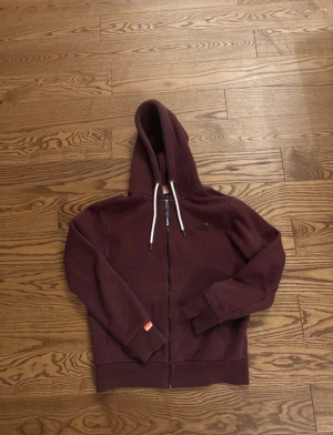 Mörk vinröd zip hoodie från Superdry - Säljer en mörk vinröd hoodie från Superdry med dragkedja och huva med vita snören. Tröjan har broderad logga på bröstet, två fickor fram och mjukt fleecefoder på insidan. Perfekt för chill dagar och har en normal passform. Något liten i storleken.