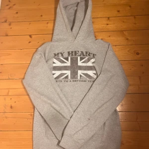 Grå hoodie med brittisk flagga - Säljer en grå hoodie med tryck på bröstet som visar en sliten brittisk flagga och texten 'MY HEART BEATS TO A BRITISH TUNE'. Tröjan har huva med snörning och långa ärmar, perfekt för dig som gillar streetstyle och coola prints.