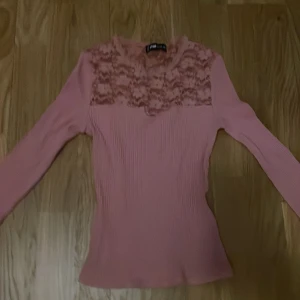 Rosa ribbad topp med spets från Rue Paris - Snygg ljusrosa långärmad topp från Rue Paris med ribbad struktur och spetsdetaljer över bröstet och axlarna. Spetsen har blommigt mönster. Perfekt att styla med jeans eller kjol för en trendig look.