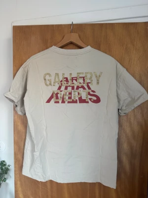 Beige Gallery Dept t-shirt med tryck - Säljer en beige t-shirt från Gallery Dept med stort tryck på ryggen i glitterguld och rött, samt ett statement-tryck på framsidan i svart och rött. T-shirten är i 100% bomull och har klassisk passform med korta ärmar och rund hals. Perfekt för dig som gillar streetwear och unika prints. Sitter som M/L!