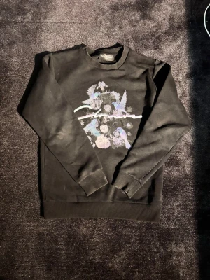 Svart sweatshirt med fågeltryck Closure - Svart sweatshirt från Closure med snyggt tryck av blå och lila fåglar samt blommor på bröstet. Tröjan har rund hals, ribbade muddar och lång ärm. Perfekt för dig som vill ha en cool och unik look.