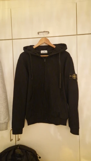 Svart hoodie från Stone Island - Svart hoodie från Stone Island med huva och snörning, dragkedja framtill och klassisk logotyp-patch på vänster ärm. Tröjan har en normal passform och är tillverkad i mjuk bomull, perfekt för chill dagar. Snygg och enkel att matcha med jeans eller joggers.