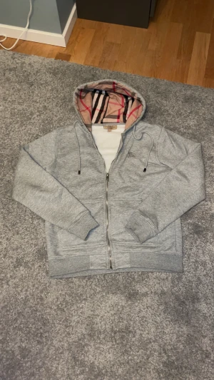 Grå Burberry hoodie med dragkedja - Snygg grå hoodie från Burberry med klassiskt rutigt foder i huvan och insidan. Tröjan har hel dragkedja, snörning vid huvan och ribbade muddar. Perfekt för dig som vill ha en stilren look med lyxig touch och ikoniskt Burberry-mönster.