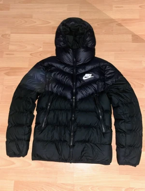 Svart Nike pufferjacka med huva - Svart pufferjacka från Nike med stor huva och vita loggan på bröstet. Jackan har två snedställda fickor med dragkedja och quiltad design. Perfekt för kalla dagar, riktigt chill och snygg stil. Materialet är syntet och jackan har normal passform.