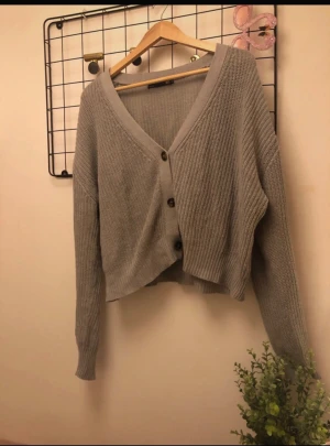 Grå croppad kofta med knappar - Snygg grå croppad kofta med v-ringning och tre knappar framtill. Koftan är stickad i ett ribbat mönster och har långa ärmar. Perfekt för lager-på-lager och chill stil. Passar dig som gillar enkel men trendig look.