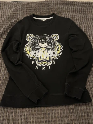 Svart Kenzo  - Svart sweatshirt från Kenzo i storlek XS med ikonisk broderad tiger och logga på bröstet. Tröjan har rund hals, ribbade muddar och lång ärm. Broderiet har detaljer i vitt, gult och blått. Perfekt för dig som gillar statement-plagg och streetstyle.