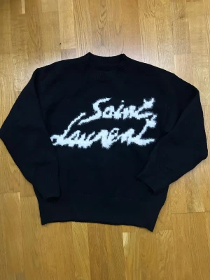 Svart stickad Saint Laurent tröja - Svart stickad tröja från Saint Laurent med vit fuzzy logga framtill. Tröjan har rund hals, ribbade muddar och en loose passform som ger en chill vibe. Perfekt för dig som vill ha något snyggt och bekvämt med designerfeeling.