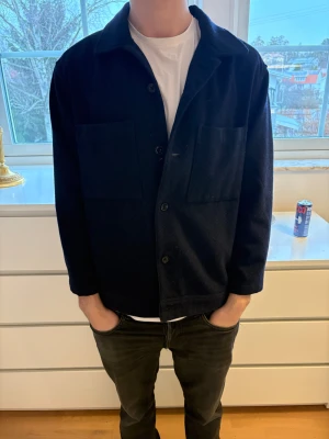 Svart overshirt från Riley i ull - Säljer en stilren svart overshirt från Riley. Jackan har två stora bröstfickor, klassisk krage och knäppning framtill. Tillverkad i mjuk ull som ger en clean och modern look. Perfekt att slänga över en t-shirt för en avslappnad vibe.