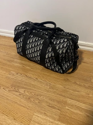 Väska dior - Säljer en snygg resväska från Dior med svart och vit mönstrad design. Väskan har både handtag och axelrem, detaljer i svart skinn och dragkedja upptill. Perfekt storlek för weekendresor och har en cool, modern look med Dior-logga över hela väskan. (Vet ej om den är äkta)