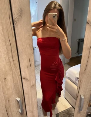 Röd axelbandslös klänning med volang - Säljer en snygg röd axelbandslös klänning med figurnära passform och volangdetalj längs sidan. Klänningen har en asymmetrisk slits och är i ett mjukt, stretchigt material som sitter bekvämt. 