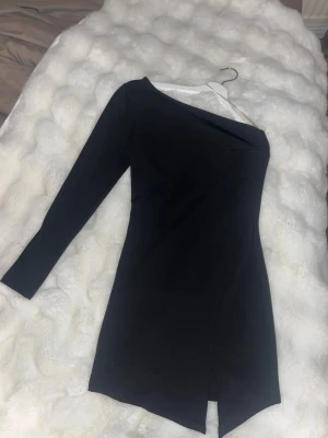 Svart oneshoulder klänning Chiquelle - Snygg svart kortklänning från Chiquelle med oneshoulder-design och lång ärm. Klänningen har en stilren, tight passform och en liten slits framtill. Perfekt för dig som gillar minimalistisk och trendig stil. Klänningen ser ut som ny och är använd en gång.