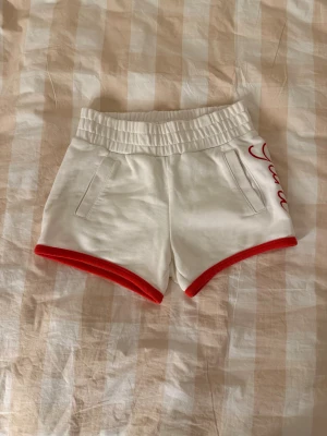Vita shorts med röd kant från Gant - Snygga vita shorts från Gant med bred resår i midjan och röda kanter längs bensluten. På ena sidan finns Gant-broderi i rött. Shortsen har två fickor fram och är gjorda i mjuk bomull, perfekta för chill dagar och sport. 