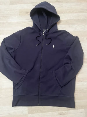 Mörkblå hoodie från Polo Ralph Lauren - Snygg mörkblå hoodie från Polo Ralph Lauren med klassisk logga broderad på bröstet. Tröjan har huva med snörning, dragkedja framtill och två fickor. Tillverkad i mjuk bomull. Perfekt för chill dagar eller för stilren look storlek M hör av er vid frågor☺️