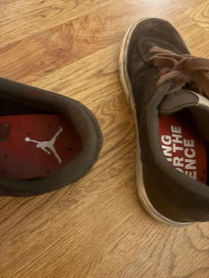 Jordan Series bruna sneakers - Säljer ett par bruna Jordan Series sneakers från Nike med bruna snören och vit sula. Skorna har röd innersula med Jumpman-loggan och textdetaljer på sidan. Ovandelen är i mocka och har en klassisk rund tå och platt sula.