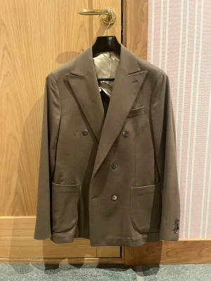 Brun dubbelknäppt kavaj Loro Piana - Snygg brun dubbelknäppt kavaj med material från Loro Piana, tillverkad i 100% ren ull från Italien (doeskin). Klassisk design med breda slag, stora framfickor och knappar vid ärmslut. Perfekt för dig som gillar stilrena och tidlösa plagg.