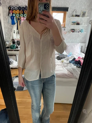 Ljusbeige henley från KappAhl - Luftig och tunn ljusbeige henley-skjorta från KappAhl med v-ringning och knappar framtill. Skjortan har trekvartsärmar med lätt rynk vid ärmslut och är tillverkad i ett mjukt bomullstyg. Själv har denna varit väldigt praktisk att ha med till stranden!