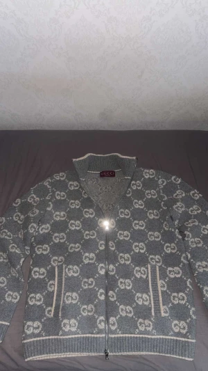 Grå cardigan jacka från Gucci - Säljer en grå cardigan jacka från Gucci med hel dragkedja och ribbad krage. Jackan har ett mönster med vita GG-loggor över hela plagget och vita detaljer vid fickor och muddar. Materialet är stickad ull som känns både lyxigt och mjukt. Storlek L