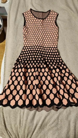 Kort ärmlös klänning med hexagonmönster - Säljer en cool kort klänning med ärmlös design och rund hals. Klänningen har ett unikt hexagonmönster i svart och beige som ger en modern vibe. Kjolen är utsvängd och materialet känns mjukt och stretchigt, perfekt för dig som gillar att sticka ut.