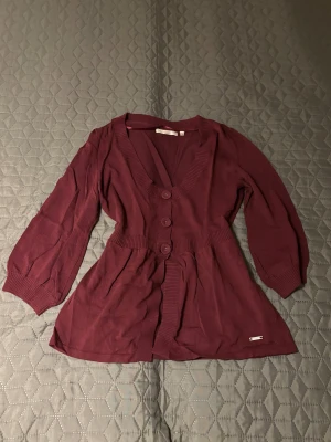 Vinröd kofta från Riley, storlek M - Snygg vinröd kofta från Riley med markerad midja och tre stora knappar framtill. Koftan har långärmade puffärmar och ribbade muddar samt v-ringad hals. Materialet är mjukt och stickat, perfekt för att ge en chill och stilren vibe.