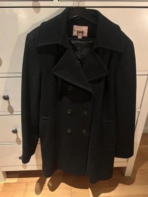 Svart skepparkavaj från Joy, stl 38 - Säljer en svart skepparkavaj från Joy Outerwear i storlek 38. Jackan har dubbelknäppning med sex knappar, bred krage och två fickor framtill. Materialet är ullblandning och modellen är klassisk med lång ärm och snygg passform.