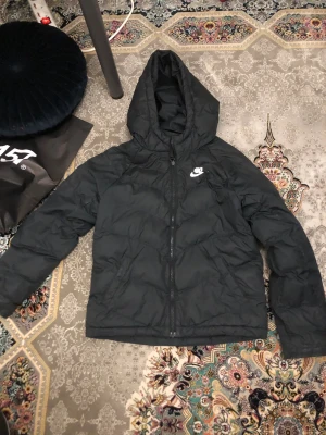 Svart pufferjacka från Nike,  - Svart pufferjacka från Nike med huva och dragkedja framtill. Jackan har ett stilrent Nike-logotryck på bröstet och quiltad design som ger extra värme. Perfekt för kyliga dagar och enkel att matcha med din streetwear. Snygg och funktionell look.