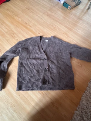 Brun kofta från Lindex med knappar - Mysig brun kofta från Lindex med ribbad struktur och tre stora knappar framtill. Koftan har v-ringad hals och långa ärmar, perfekt för att hålla sig varm under kyliga dagar. Materialet känns mjukt och stickat, vilket gör den skön att ha på sig.