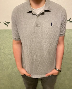 Grå pikétröja från Polo Ralph Lauren - Snygg grå pikétröja från Polo Ralph Lauren med klassisk krage och två knappar framtill. Tröjan har korta ärmar och den ikoniska mörkblå logon broderad på bröstet. Tillverkad i mjuk bomull som är skön mot huden. Perfekt för en chill och stilren look.